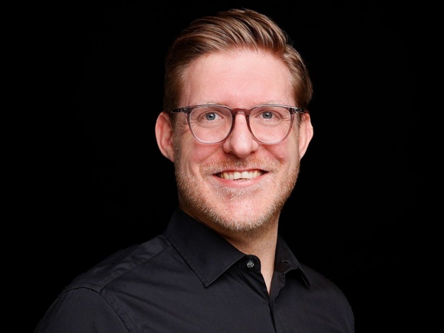 Ein hellhäutiger Mann mit Brille und Dreitagebart strahlt in die Kamera. Der Hintergrund ist schwarz.