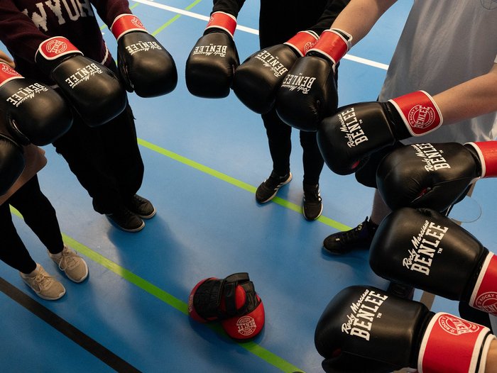 Boxhandschuhe werden im Kreis in die Mitte gehalten, man sieht nur Füße und Arme und den Boden einer Turnhalle.