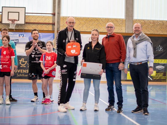 Eine Gruppe von Menschen, darunter Kinder in Sportuniformen und Erwachsene, steht während einer Preisverleihung in einer Turnhalle, ein Mann hält einen Defibrillator, eine Frau einen Spendenscheck, während sie in die Kamera schauen