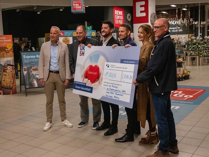 Eine Frau und fünf Männer stehen vor dem Eingang zum REWE Center und halten einen Spendencheck in den Händen.