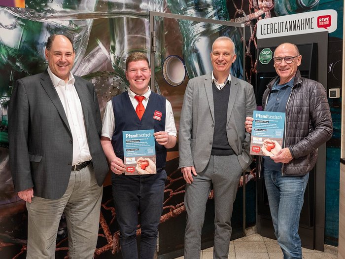 Vier Männer stehen vor einem Pfandspendeautomaten und einer bemalten Rückwand; zwei Männer halten lächelnd ein Pfandspendenplakat in den Händen.