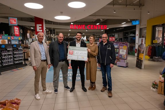 Eine Frau und vier Männer stehen vor dem Eingang zum REWE Center und halten einen Spendencheck in den Händen.
