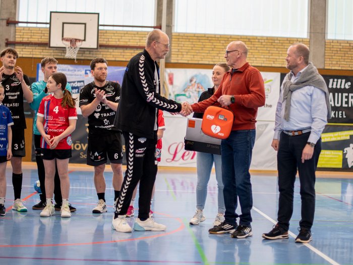 Zwei Männer schütteln sich in einer Sporthalle die Hände; Kinder und Erwachsene in Sportkleidung schauen zu; ein Mann überreicht einen Defibrillator.