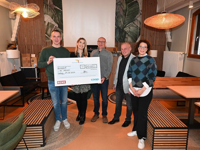 Fünf Personen stehen in einem Café ähnlichen, mit braunen Holzelementen ausgestatteten Raum. Das Licht ist warm und die Personen halten einen großen Spendenscheck über 10.000 € in den Händen.