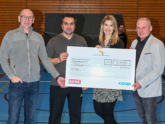 Vier Personen stehen in einer Turnhalle und halten einen großen Spendenscheck über 3.000 € in die Kamera. Im Hintergrund wärmen sich Jugendliche mit Seilspringen auf.