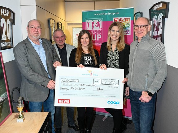 Fünf Personen stehen eng in einem Flur mit einem großen Spendenscheck über 12.000 € in der Mitte. Im Hintergrund sieht man ein Rollup auf dem das Friendscup e.V. Logo zu erkennen ist.