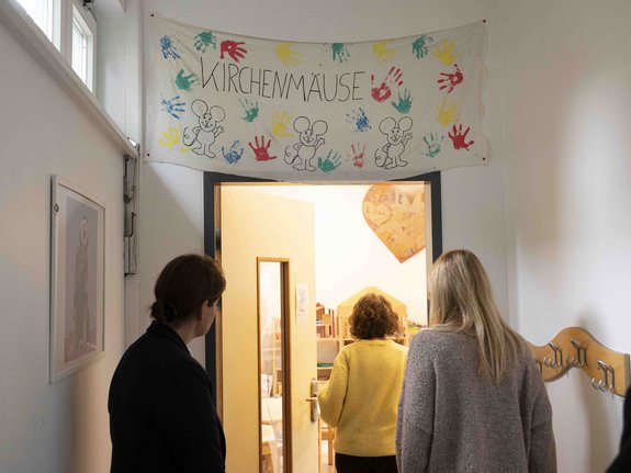 Drei Personen betrete einen Raum der Arche, über dessen Eingang ein buntes, mit Handabdrücken und Mäusen gestaltetes Banner mit der Aufschrift "Kirchenmäuse" hängt.