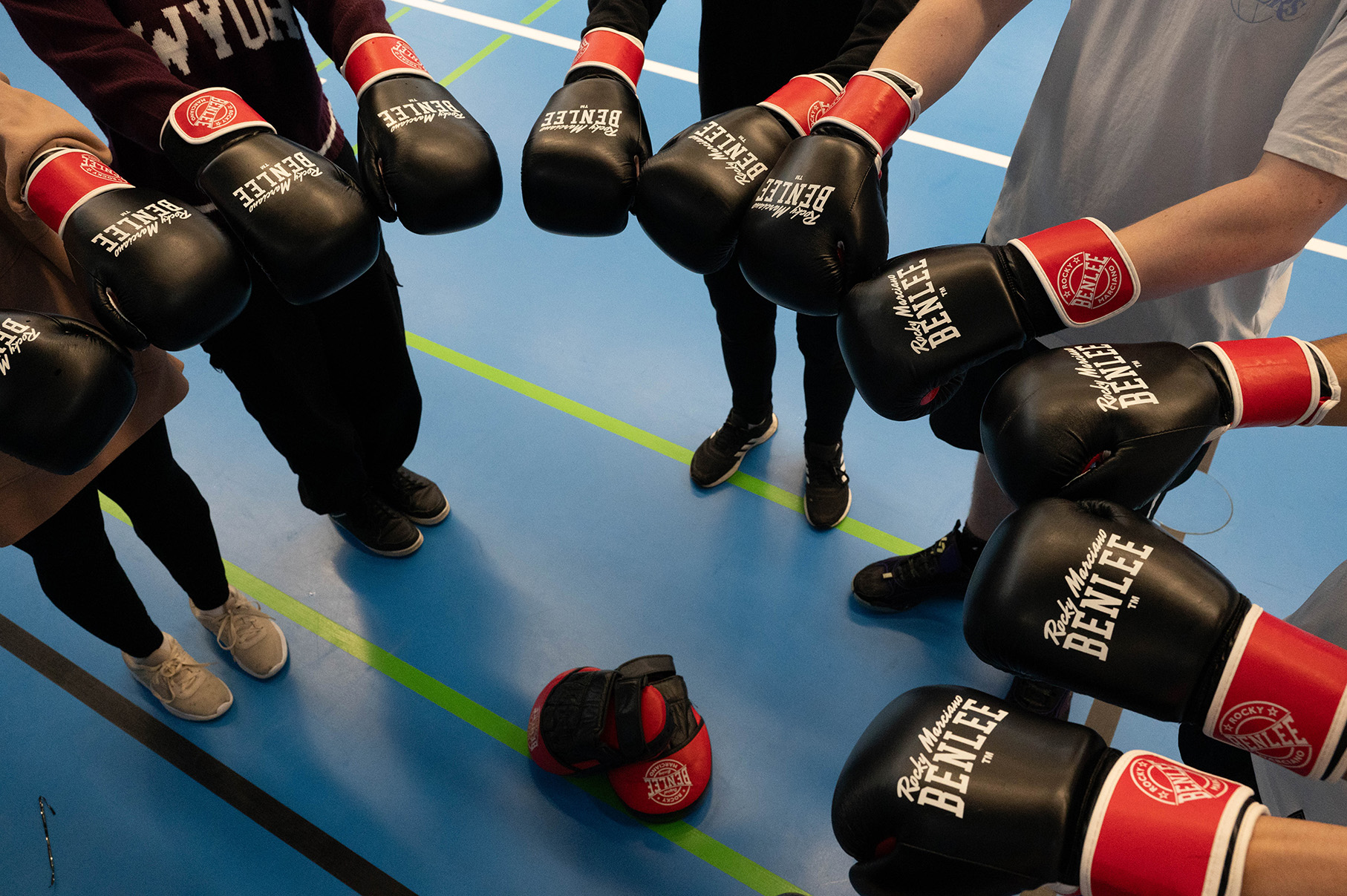 Boxhandschuhe werden im Kreis in die Mitte gehalten, man sieht nur Füße und Arme und den Boden einer Turnhalle.