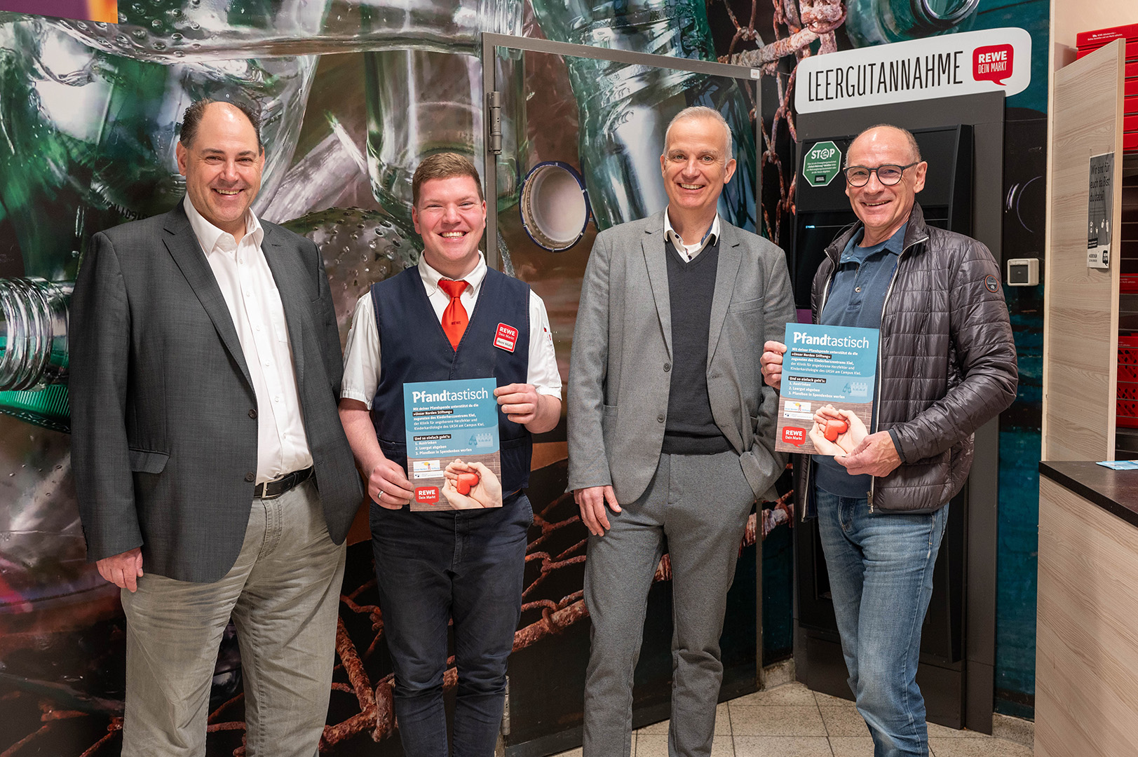 Vier Männer stehen vor einem Pfandspendeautomaten und einer bemalten Rückwand; zwei Männer halten lächelnd ein Pfandspendenplakat in den Händen.