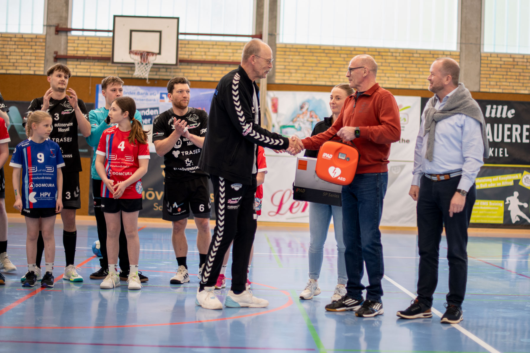 Zwei Männer schütteln sich in einer Sporthalle die Hände; Kinder und Erwachsene in Sportkleidung schauen zu; ein Mann überreicht einen Defibrillator.