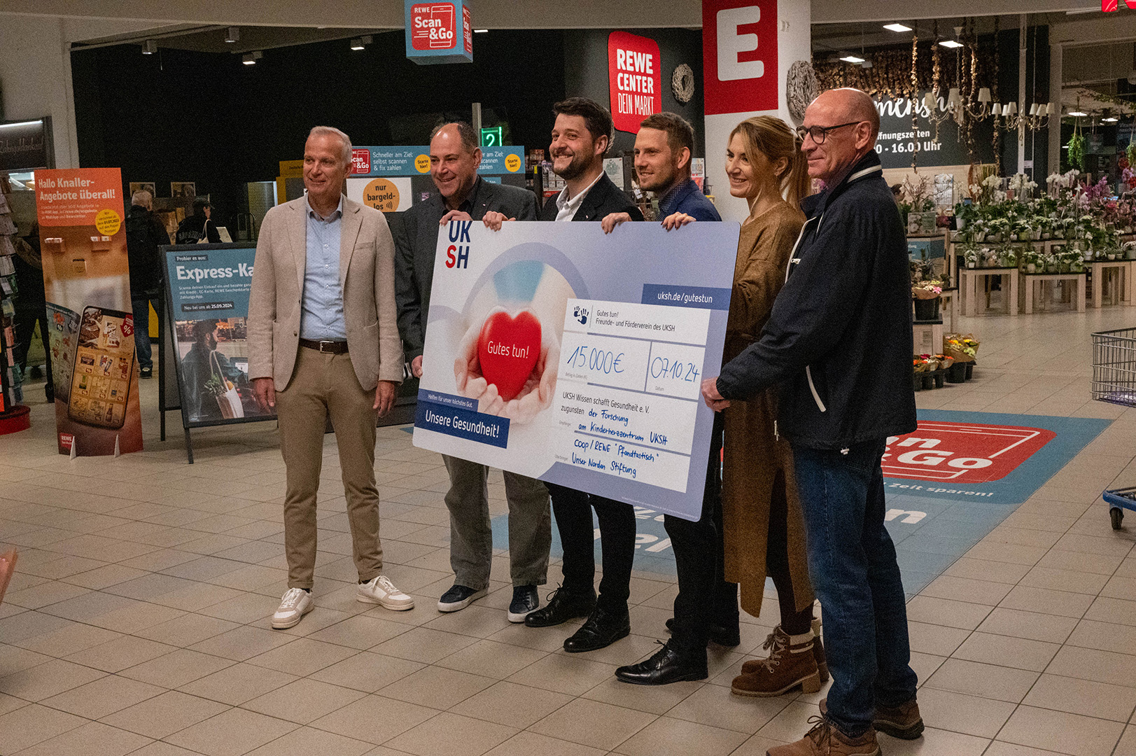 Eine Frau und fünf Männer stehen vor dem Eingang zum REWE Center und halten einen Spendencheck in den Händen.